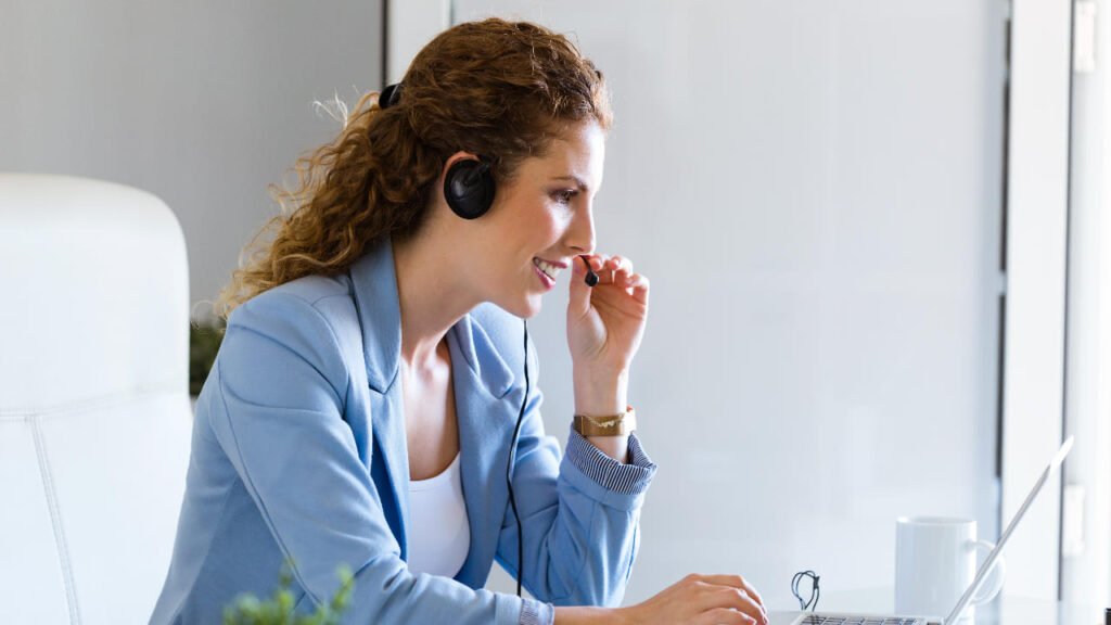 Vrouwelijke klantenservice medewerker met headset werkt op laptop. Professionele dame met koptelefoon biedt online ondersteuning. Lachende vrouw in blauw jasje met headset achter laptop. Helpdesk medewerker in gesprek met klant via computer. Zakenvrouw met headset typt op laptop voor klantenservice.