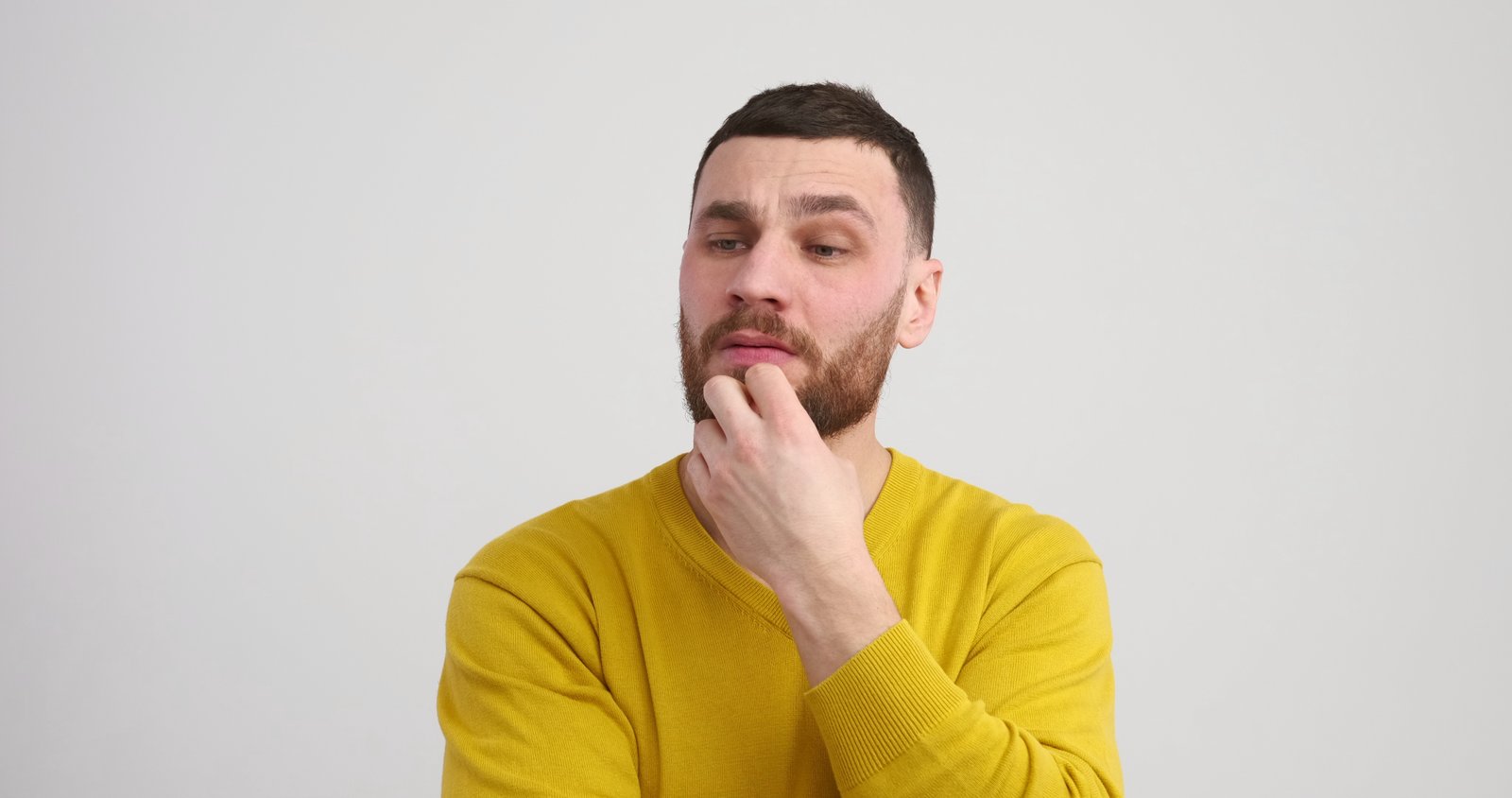 Nadenkende jonge man met baard in een gele trui, kijkt opzij met een hand aan zijn kin.